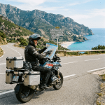 Motorcyclist sitting on a bike, holding a map by a road sign with coastal mountains and a beach in the background